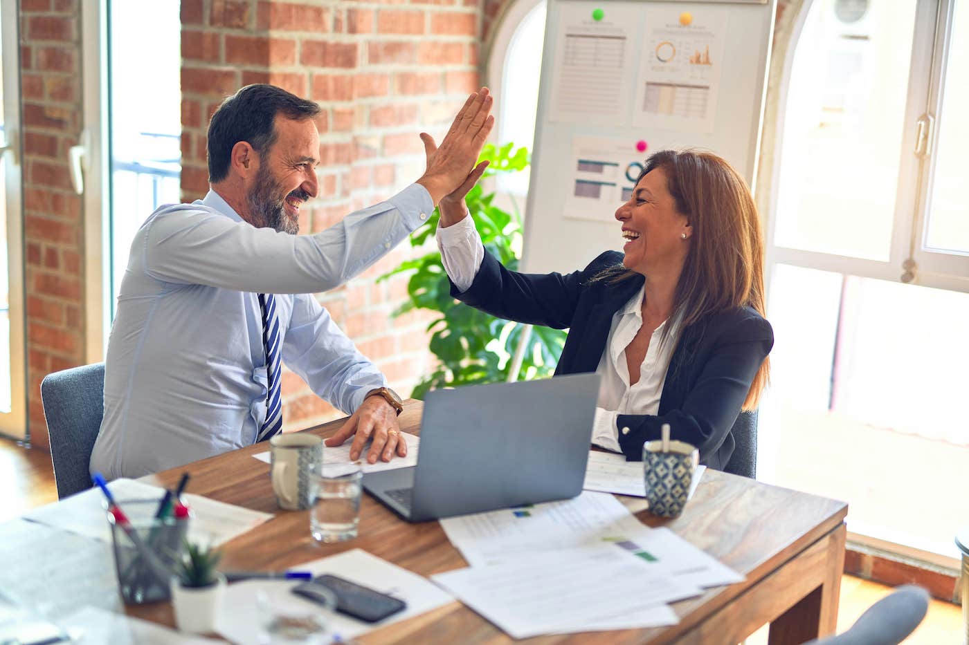 Two business colleagues high-fiving each other in an office, celebrating success with a laptop and documents on the desk.